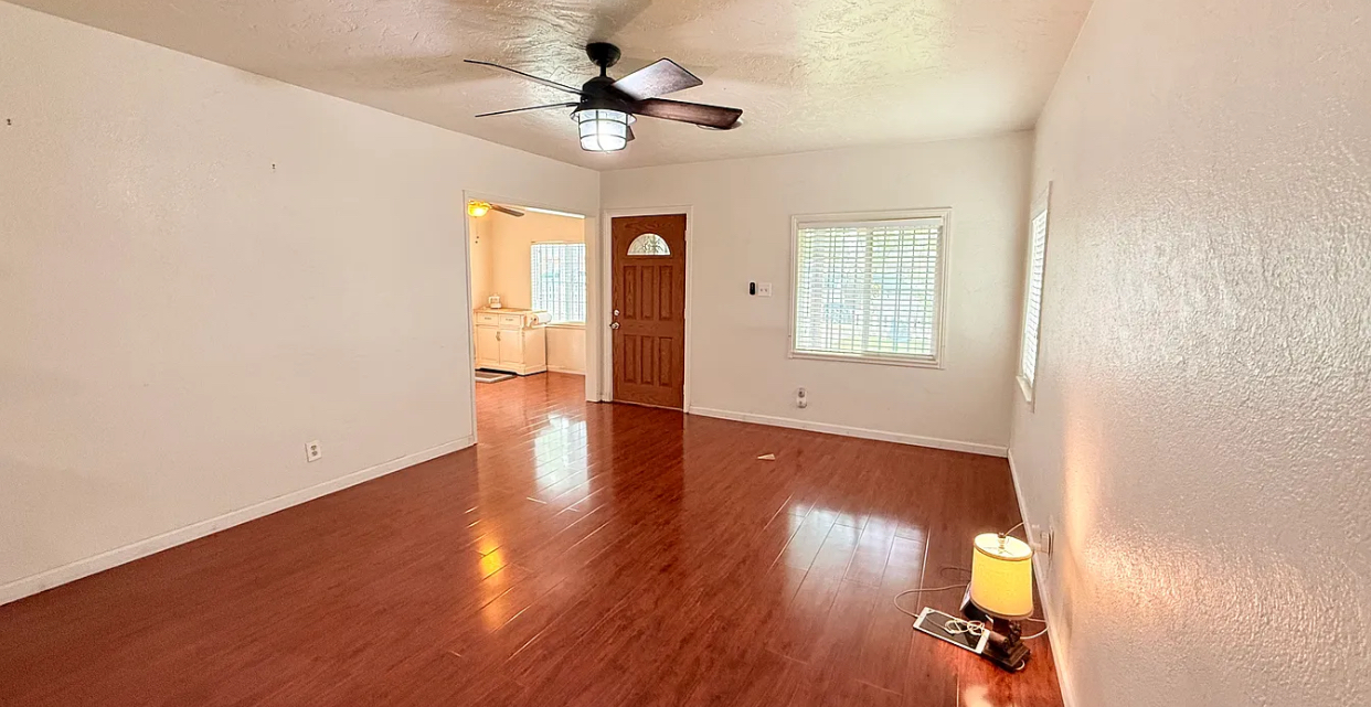 This one-bedroom apartment offers a spacious and inviting living area with ample natural light streaming in from the large window. The light grey walls and white door create a clean and modern aesthetic, while the light brown laminate flooring adds warmth and comfort. The living room is currently unfurnished, providing a blank canvas for your personal style and preferences. The doorway leads to a separate room, likely a kitchen, where a white refrigerator can be seen, suggesting a well-equipped space for preparing meals. Overall, this apartment presents a blank slate for creating a cozy and functional home, with the potential to be customized to your unique taste.